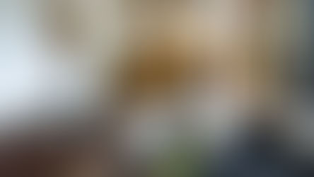 Modern living room with a black sofa, patterned wood sculpture, and plants. Woven chairs around a wooden table under pendant lights. Cozy and stylish.