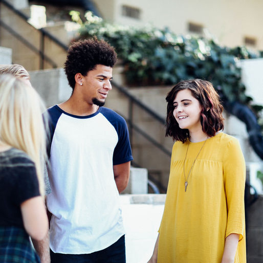 Four young adults are talking outside near stone steps and greenery. One wears a yellow dress, another a white shirt. They appear happy.