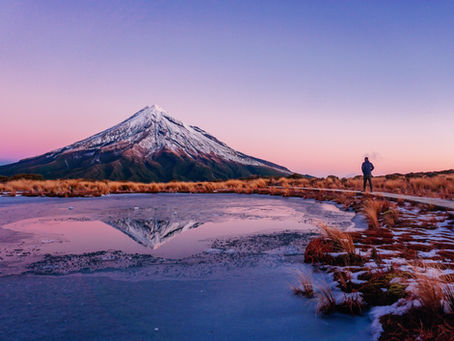 Person standing by a frozen lake reflecting snow-capped Mount Taranaki at sunset. Sky is pink and purple, creating a tranquil mood.