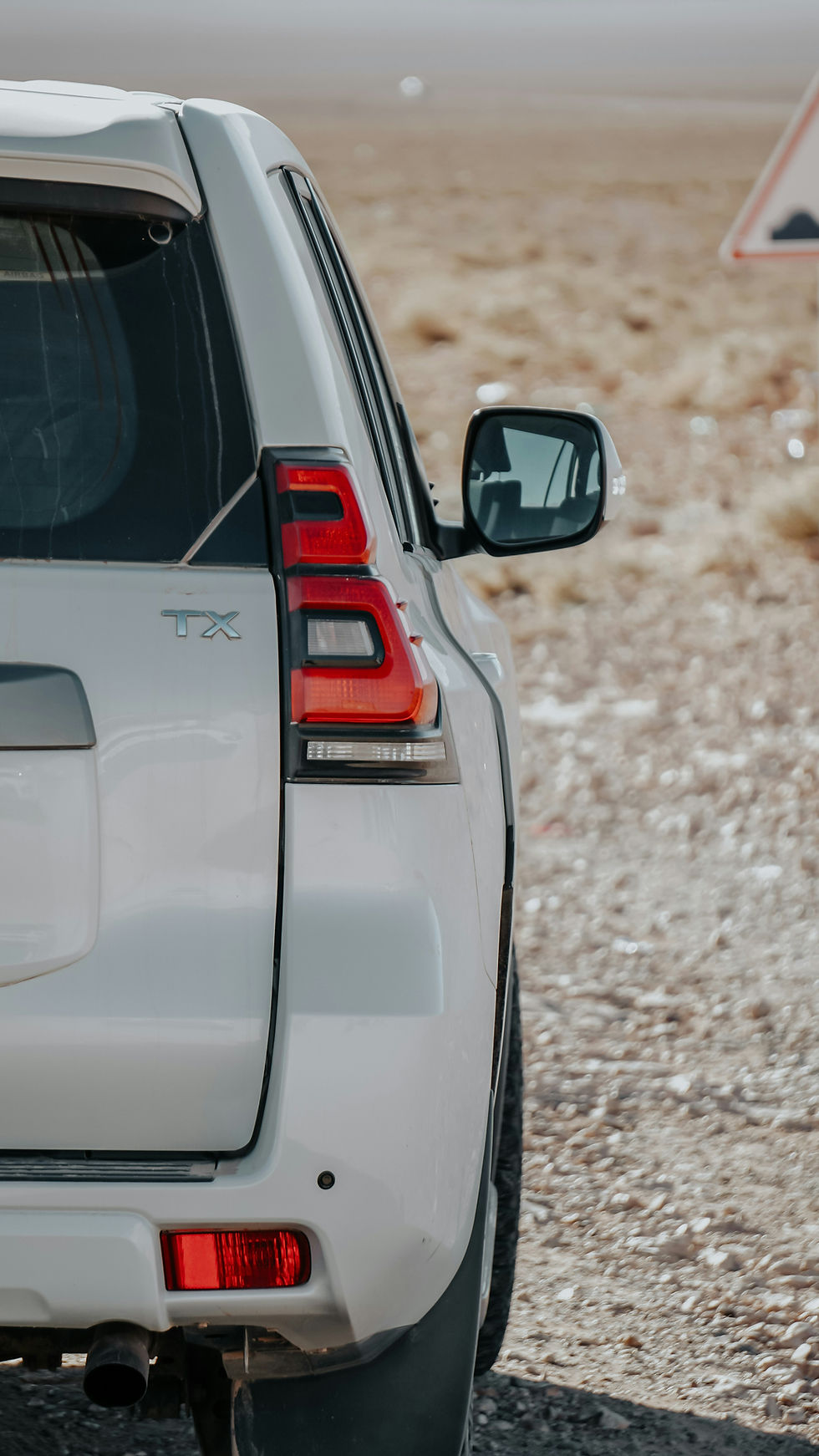 White SUV on a rocky desert road, rear side close-up with TX badge and red taillights; blurred warning sign in background.