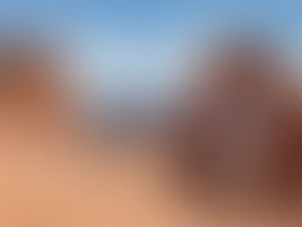 People exploring Delicate Arch in Utah's red rock landscape. Snow-capped mountains in the distance, blue sky with clouds above.