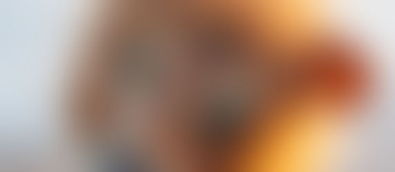 Person wearing a realistic lion mask outdoors, gazing softly at the camera. The mane is shaggy and bright against a blurred, sunny backdrop.