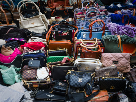 Rows of colorful luxury handbags and totes, with visible brand logos like Louis Vuitton, Prada, Dior and Bimba, filling a market stall