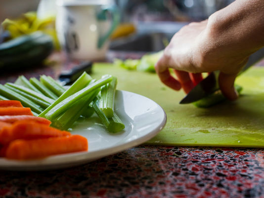 dos manos que estan cortando verdura con una cuchilla, y al lado hay un plato balnco con pencas de acelga y zanahorias peladas