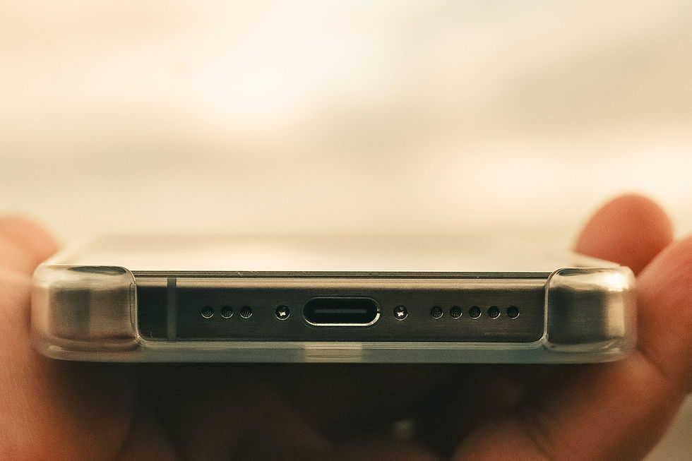 Close-up of a smartphone's charging port in a clear case, held by fingertips. Background is blurred with warm tones.