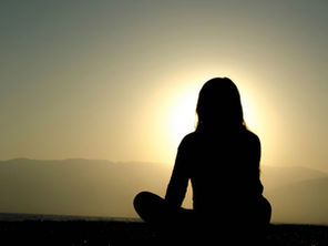 Silhouette of a person meditating at sunset, seated cross-legged on a beach. Sunlight creates a serene glow against the mountains.