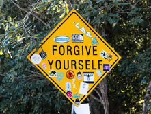 Yellow sign with "Forgive Yourself" text and stickers, set in a wooded area; evokes a reflective mood.