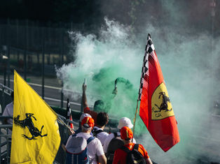 Fans on a racetrack bridge hold yellow and red flags in smoke. Energetic mood, surrounded by a green haze, with trees in the background.