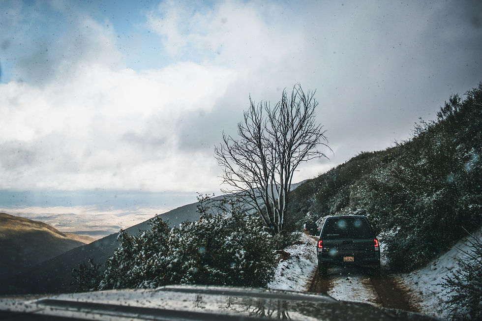 A silhouetted car drives down a snowy, winding road surrounded by bare trees and mountainous terrain.