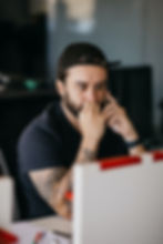 Man with tattoos in a cap, looking worried while on the phone. He's seated at a desk with a blurred office background.