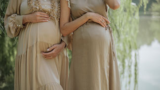 Two pregnant women in beige dresses stand outdoors by a lake, hands on their bellies, in a calm, soft-lit scene.