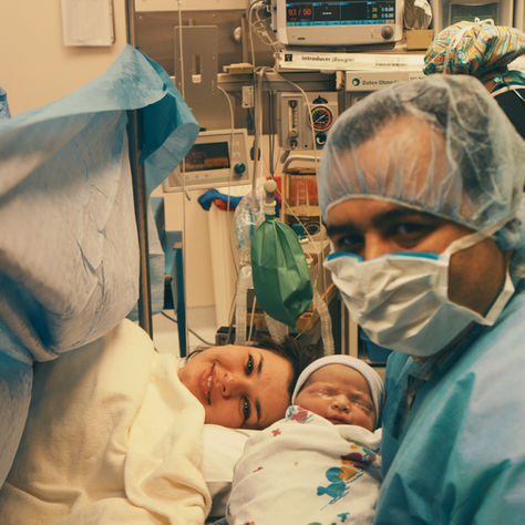 Newborn baby with mother and masked medical staff in a hospital room. The baby is wrapped in a patterned blanket, creating a warm scene.