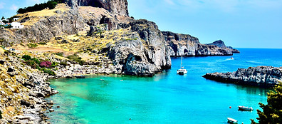 Rocky cliffs surround a turquoise bay with clear water. A white sailboat is anchored in the bay, and a few small boats float nearby. The landscape is sunlit, with sparse greenery and a bright blue sky overhead.
