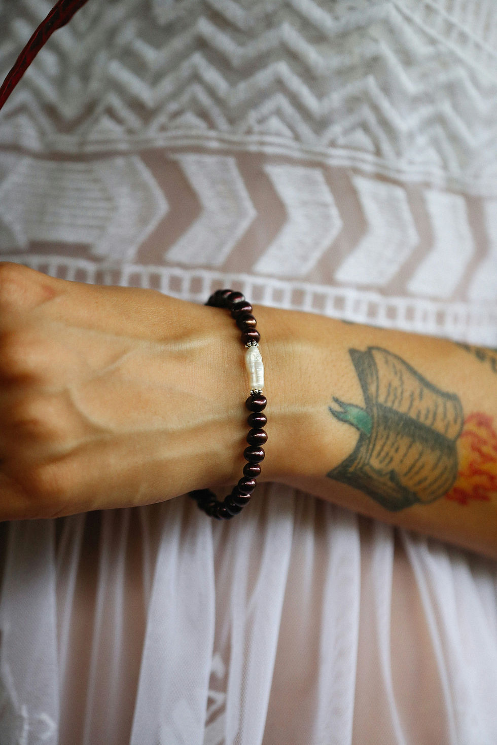 Tattooed arm wearing a dark beaded bracelet with a white centerpiece, against a patterned white fabric background.