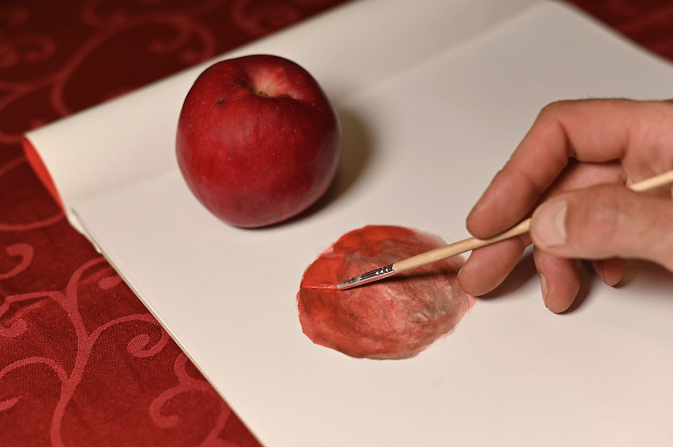 Hand painting a red apple on white paper beside a real apple, on a red patterned tablecloth.