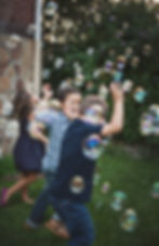 Children joyfully chase bubbles in a grassy yard, surrounded by trees. They wear colorful summer clothes, creating a playful, vibrant scene embodying ACT principles of engaging in meaningful activities that results in intrinsic motivation and in turn aligning with goals and new Year's resolution's.