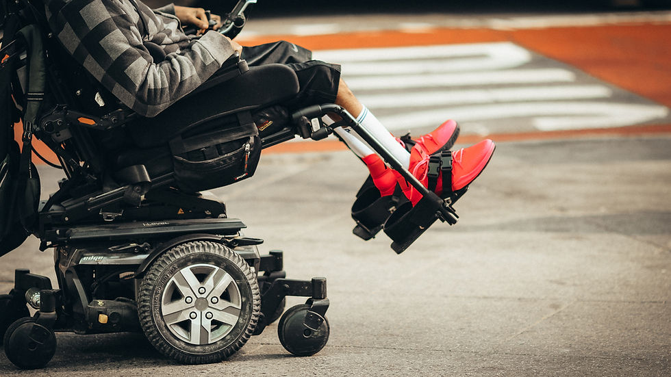 Person in a power wheelchair on a city street, wearing bright red shoes. Gray and black checkered hoodie. Urban crosswalk in background.