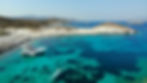 A yacht is anchored in clear turquoise waters near rocky shores. People swim nearby, enjoying the sunny day. Mountains are visible in the distance.