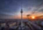 Berlin TV Tower rises over the city skyline at sunset, with orange clouds and a dramatic evening sky.