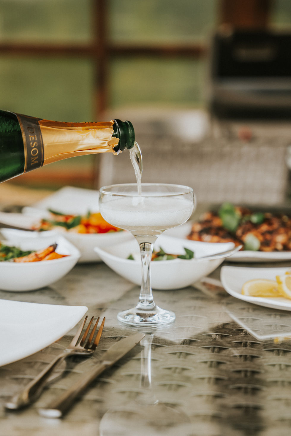 Bottle of sparkling wine being poured into a coupe glass on a table set with small white bowls of appetizers