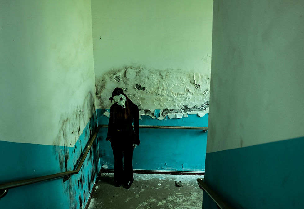 Masked figure in black stands in a moldy, peeling stairwell with turquoise walls, creating a eerie, abandoned mood.