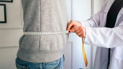 Doctor in white coat measures a person’s waist with a tape measure in a bright clinic, focused health check.