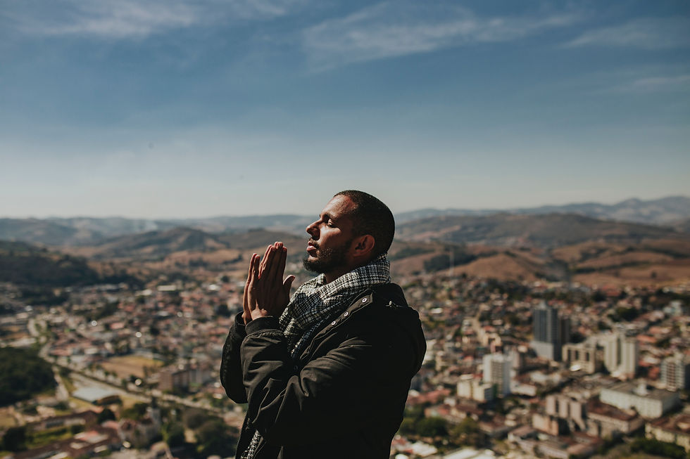 Man stands outdoors with hands clasped in prayer, eyes closed, overlooking a city with rolling hills under a clear blue sky. Calm mood.