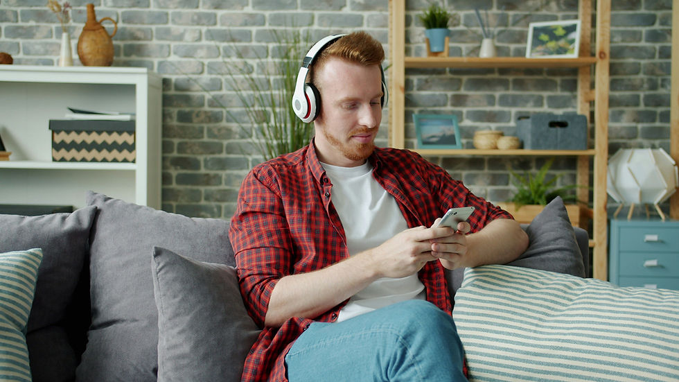 Man in headphones, red plaid shirt, and jeans using a smartphone on a gray sofa. Brick wall, shelves, and plants in the background. Cozy mood.