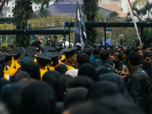 Graduation ceremony with students in yellow gowns and black caps, surrounded by a crowd. Trees and buildings visible in the background.
