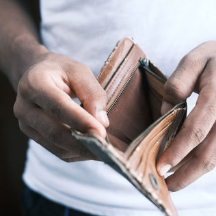 Hands holding an open, empty brown wallet. Person wearing a white shirt. The mood is contemplative or concerned. No text visible.