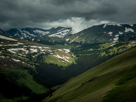 mountains speckled with snow and clouds with green hills and pines in front of it.