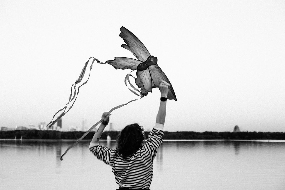 Person in striped shirt flying a bird-shaped kite by a calm lake, with a city skyline in the background. Black and white image.