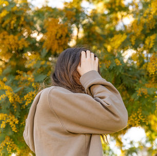 Person in a brown hoodie holds their head with hand, standing outdoors. Background features lush green foliage with yellow flowers.