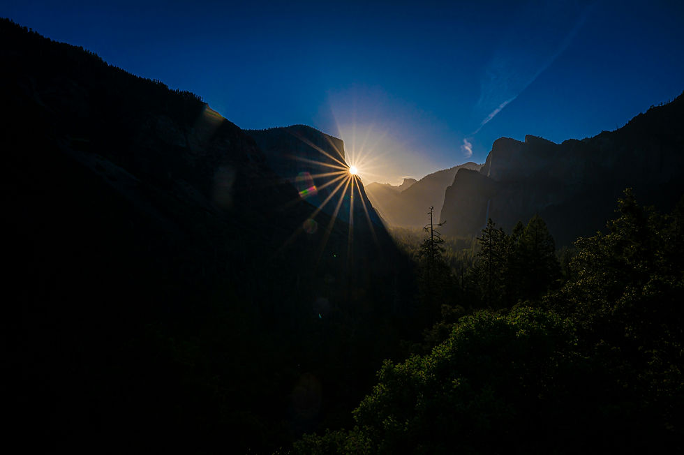 Sunrise starburst over a forested mountain valley, with dark cliffs silhouetted against a deep blue sky and misty light