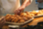 Hands reach for croissants on a wooden counter. Trays of golden pastries fill the scene, creating a warm, inviting atmosphere.