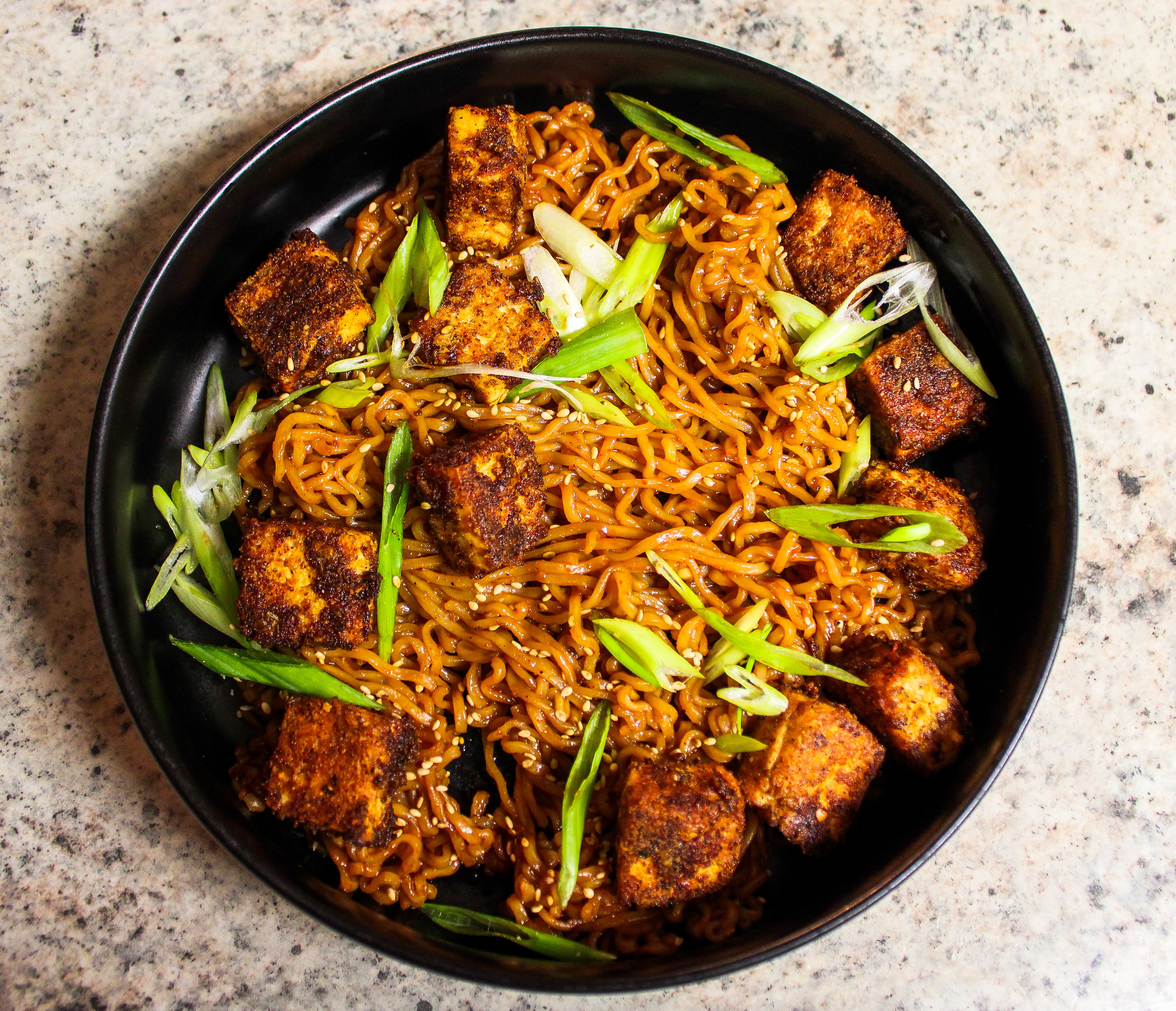 A black bowl of spicy tofu stir-fry with noodles, garnished with green onions and sesame seeds, on a speckled countertop.
