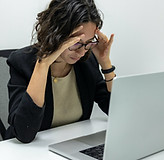 Exhausted woman at a computer holding her forehead, illustrating burnout and chronic stress, representing women seeking burnout therapy in Illinois and online support for nervous system recovery and balance.
