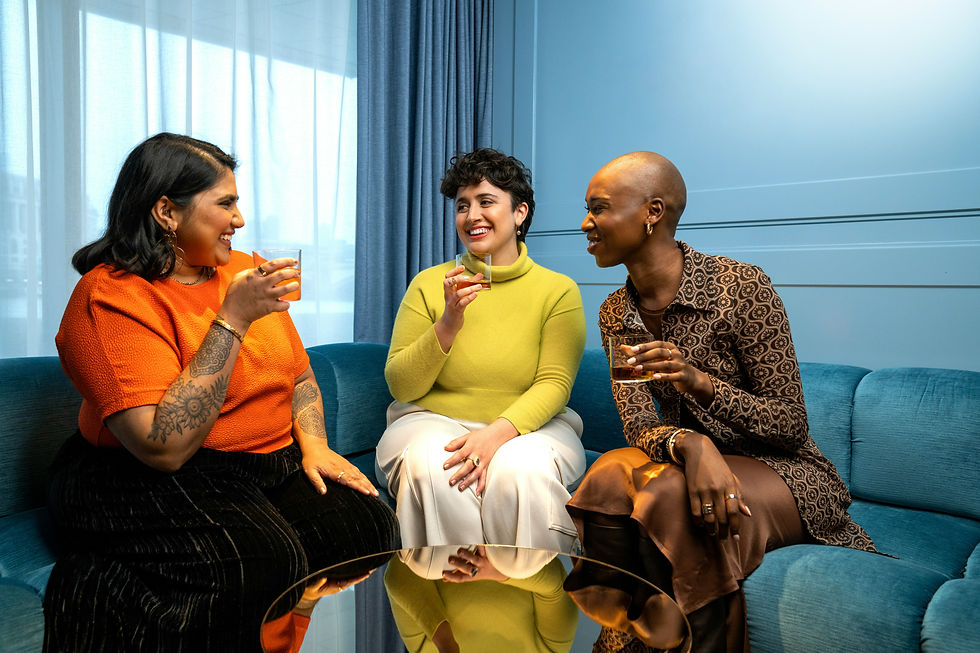 Three women smile and chat with drinks on a blue couch in a bright modern room.