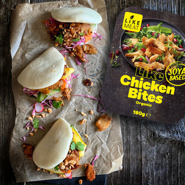 Three bao buns with soy chicken bites on parchment beside a Like Meat soy-based chicken bites package on dark wood.