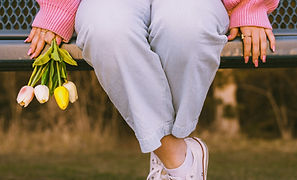 A woman wearing a pink sweater, grey trousers and white sneakers, seating on a park bench. View of only her legs and hands holding colourful tulips.