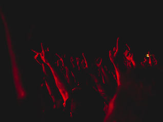 Raised arms in a dark, red-lit concert crowd creating an energetic atmosphere. Hands show rock and peace signs. No visible text.