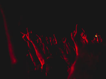 Raised arms in a dark, red-lit concert crowd creating an energetic atmosphere. Hands show rock and peace signs. No visible text.