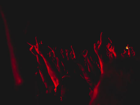 Raised arms in a dark, red-lit concert crowd creating an energetic atmosphere. Hands show rock and peace signs. No visible text.