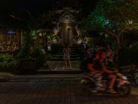 Vibrant Nusa Dua nightlife scene with a warmly lit restaurant entrance, people dining, and motorbikes passing by in Bali.