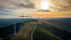 Wind turbines on a grassy hill under a cloudy sky, with a winding path and vibrant sunset in the background. Peaceful and scenic. India's Green Energy expansion