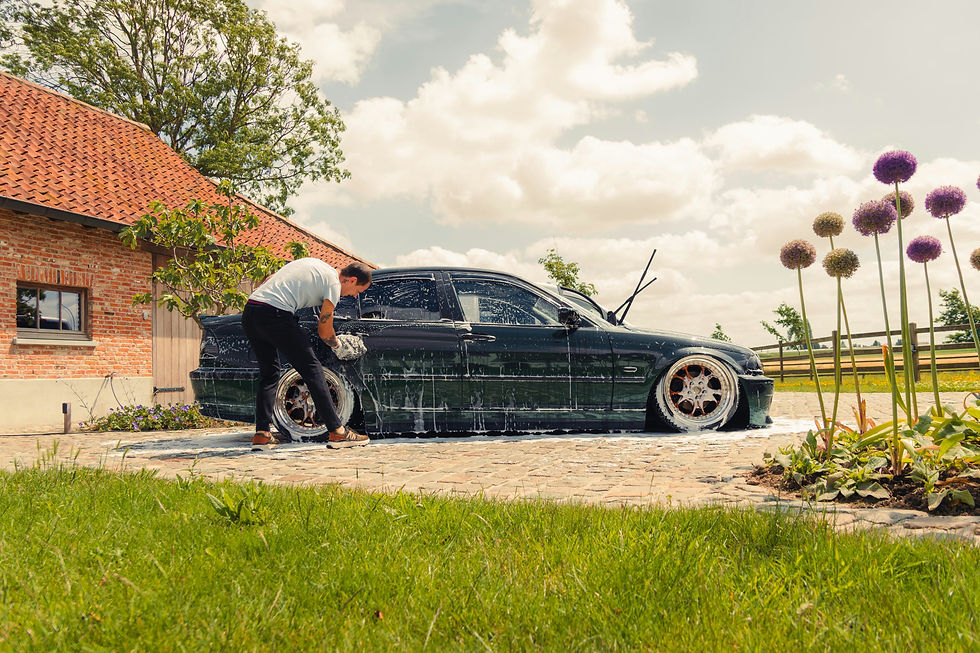 Man washing a black sedan with soap outside a brick house, beside purple flowers and green lawn under a cloudy sky.