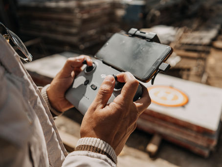 Person operates a drone controller with a smartphone attached, set outdoors in a sunny setting. Orange landing pad visible in the background.