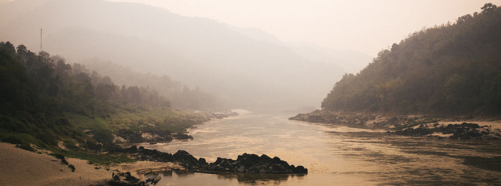 A small canoe style boat floating along a river between two hills with fog covering the land.