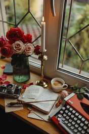 Bouquet of roses, magic wand lying across an open notebook, mug, and typewriter in front of an antique metal framed window, creating a mystical, embellished vibe.