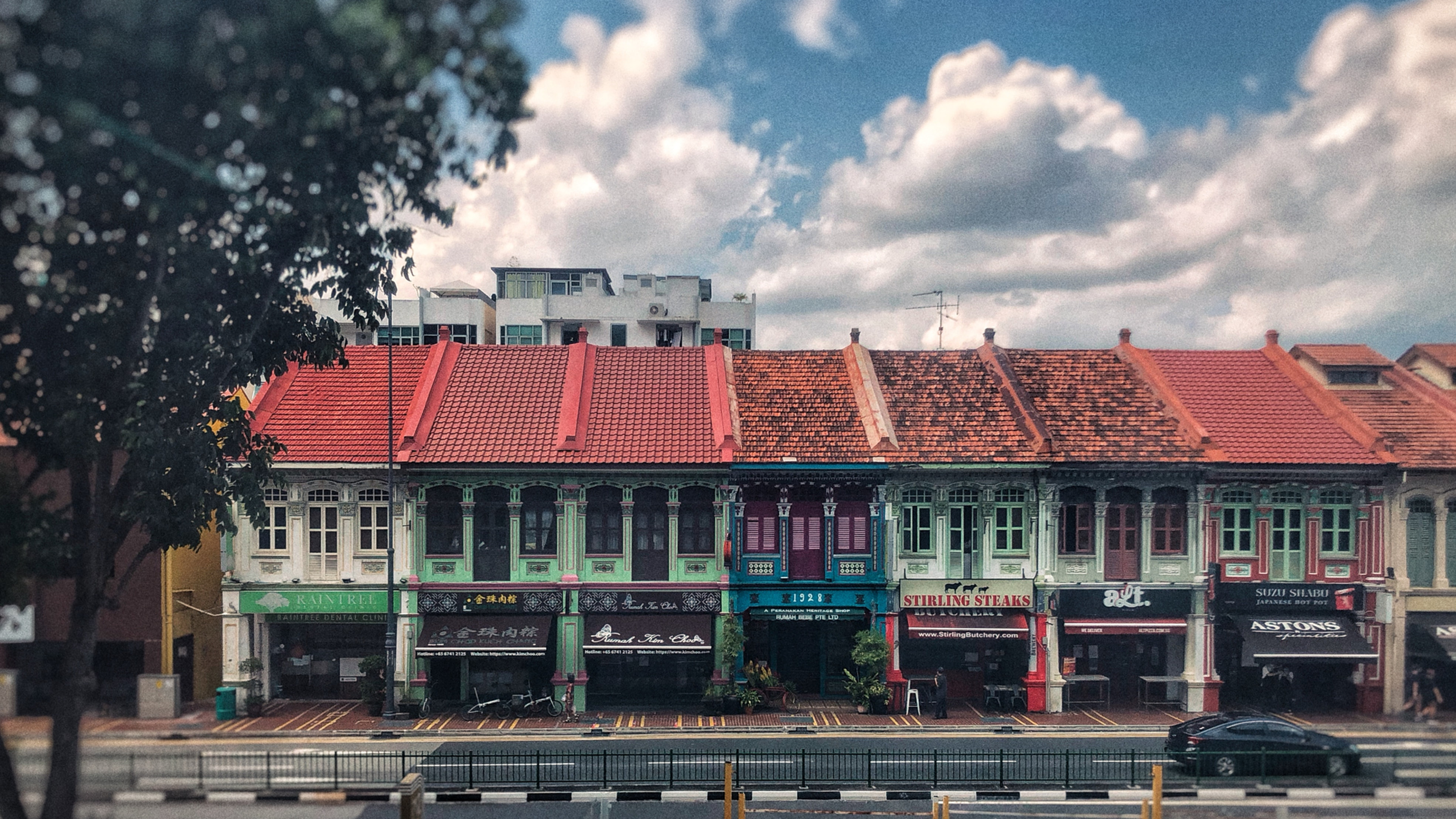 Row of colorful shophouses with red-tiled roofs and storefront signs under a bright cloudy sky, with a car passing in front